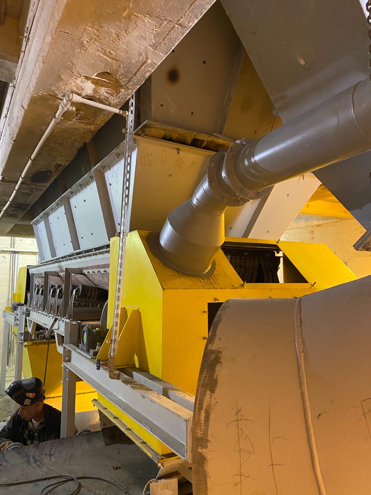 Wnętrze hali przemysłowej z fragmentem linii produkcyjnej w odcieniach szarości i żółci, spawacz w masce ochronnej przy pracy.