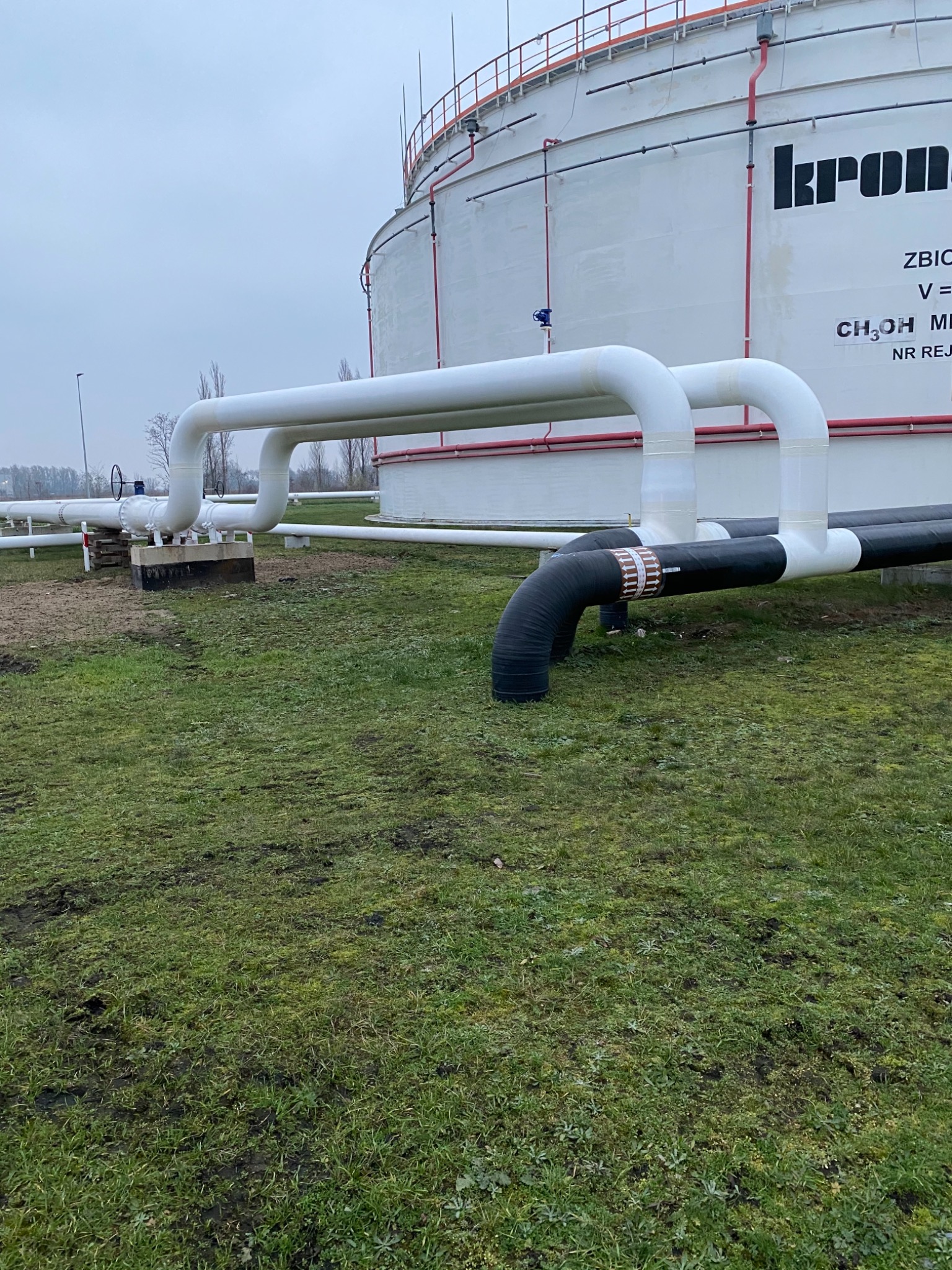Biały zbiornik przemysłowy z widocznymi rurociągami, niektóre pomalowane na biało, inne na czarno, na tle zielonej trawy i pochmurnego nieba.