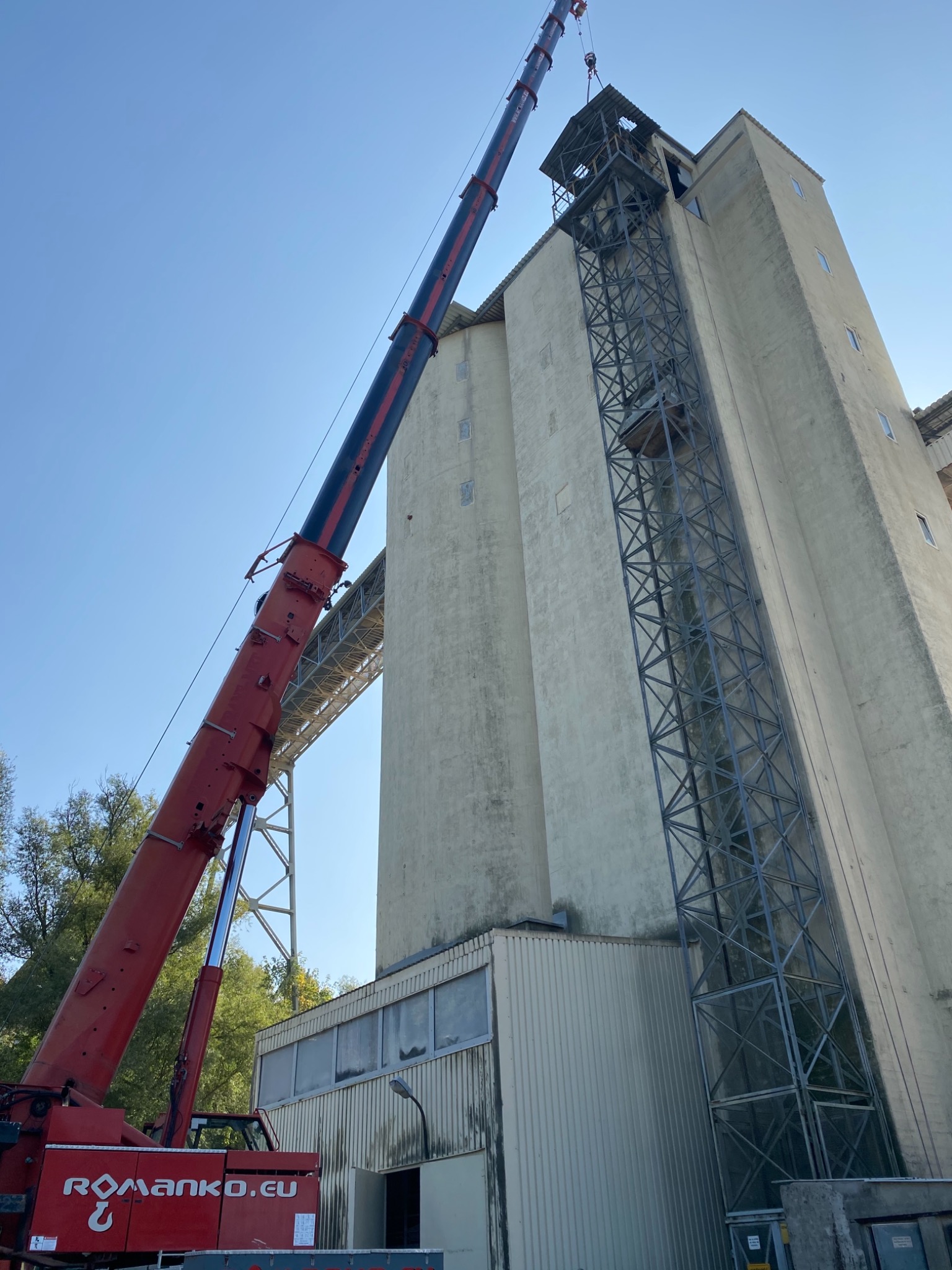 Czerwony dźwig teleskopowy z wysięgnikiem ustawiony przy wysokim, betonowym silosie z metalową konstrukcją klatki schodowej na zewnątrz.