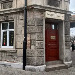 Fasada kamienicy z szyldem 'Biuro Tłumaczeń Katarzyna Sarnowska' nad czerwonymi drzwiami wejściowymi i napisem 'Biuro Tłumaczeń' w oknie, widok z ulicy wyłożonej kostką.