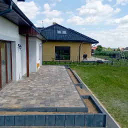 Ułożona kostka brukowa w odcieniach szarości i beżu na posesji, z obrzeżami w kolorze grafitowym, widok na dom z białymi ścianami i brązowymi oknami oraz trawnik z elementami placu zabaw w tle.