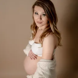 Portret kobiety w ciąży, ubrana w biały biustonosz i kremowy kardigan, z ręką na brzuchu, na jednolitym beżowym tle.