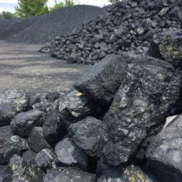 Stos czarnego, błyszczącego węgla kamiennego w składzie opału, z widocznymi złocistymi inkluzjami w niektórych bryłach, na betonowej powierzchni, z kopcem węgla w tle.