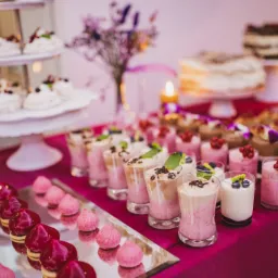 Bogato zastawiony stół z deserami: mini bezy na paterze, musy w szklankach z owocami i czekoladą, małe ciastka polane glazurą, w tle torty i bukiet kwiatów.