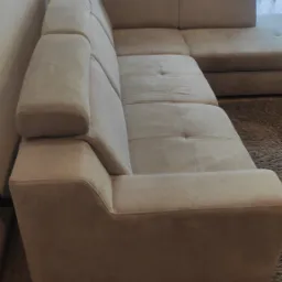 Beżowa, narożna kanapa z tapicerowanymi podłokietnikami ustawiona w rogu pokoju, z widoczną zasłoną w tle.