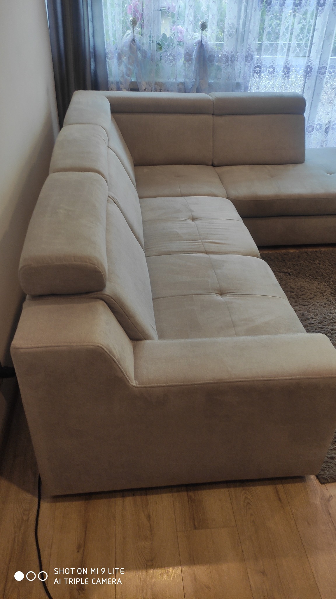 Beżowa, narożna kanapa z tapicerowanymi podłokietnikami ustawiona w rogu pokoju, z widoczną zasłoną w tle.