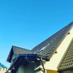 Nowy dach z czarnej dachówki ceramicznej, widoczne okno dachowe, system odgromowy i rynna PCV na tle błękitnego nieba.