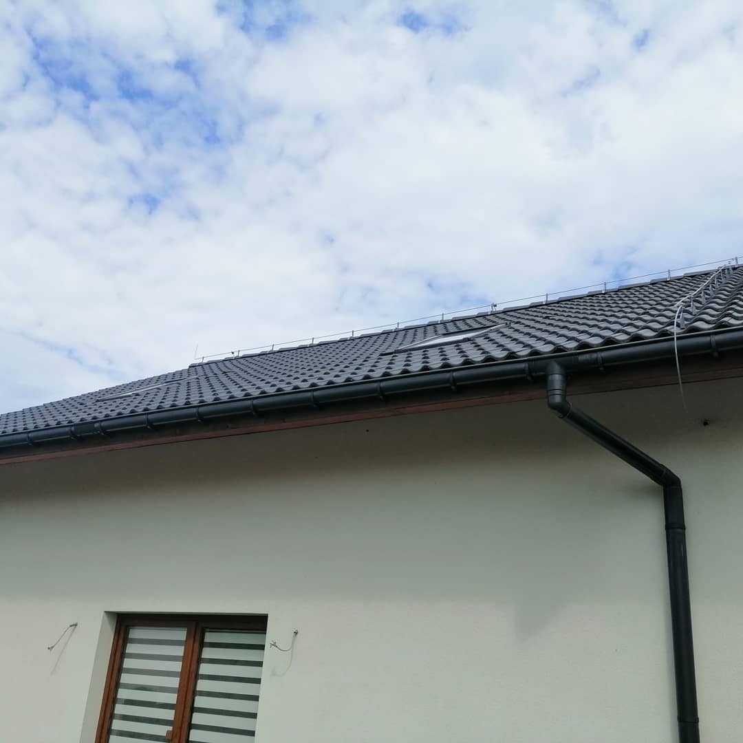 Nowy, grafitowy dach z ceramicznej dachówki, widoczna rynna spustowa i okno dachowe, na tle błękitnego nieba z chmurami.