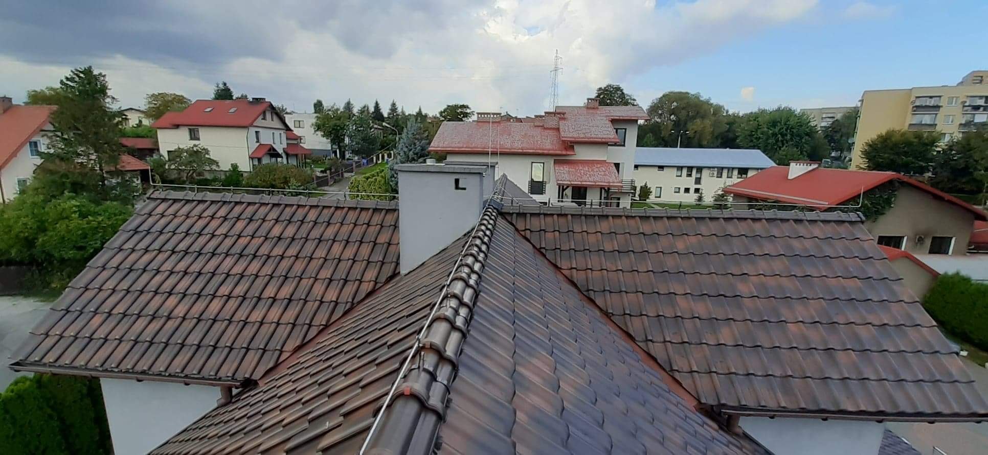 Dach z brązowej dachówki ceramicznej, widok z góry na tle zabudowy jednorodzinnej i pochmurnego nieba.