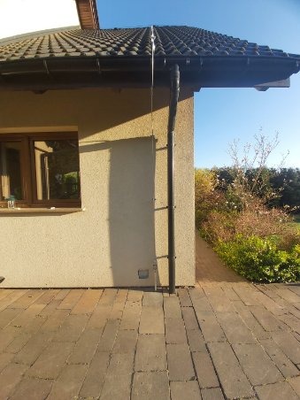 Elewacja budynku z ciemną rynną spustową, widoczne brązowe okno i dach pokryty ciemną dachówką, na pierwszym planie kostka brukowa