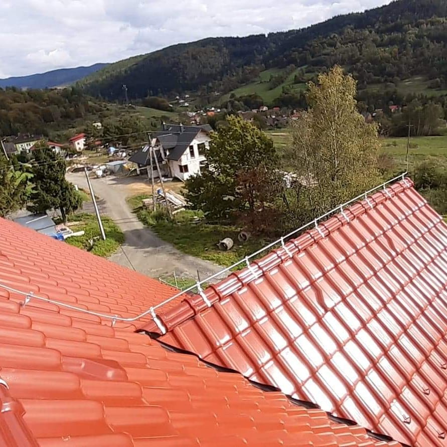 Czerwona dachówka ceramiczna z systemem odgromowym na tle górskiego krajobrazu z widocznymi zabudowaniami.