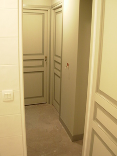 Wnętrze w trakcie remontu: widok na korytarz z dwiema odnowionymi, pomalowanymi na jasnozielono drzwiami z dekoracyjnymi panelami. Podłoga w surowym stanie, ściany przygotowane do wykończenia.