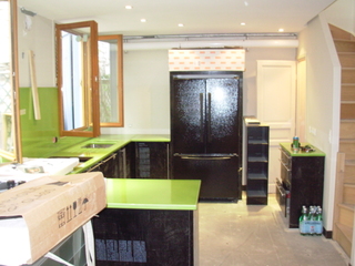 Wnętrze kuchni w trakcie remontu: czarne szafki z zielonymi blatami, otwarte okno z drewnianą ramą, czarna lodówka obłożona kartonem ochronnym, kartonowe pudło na pierwszym planie, widoczne...