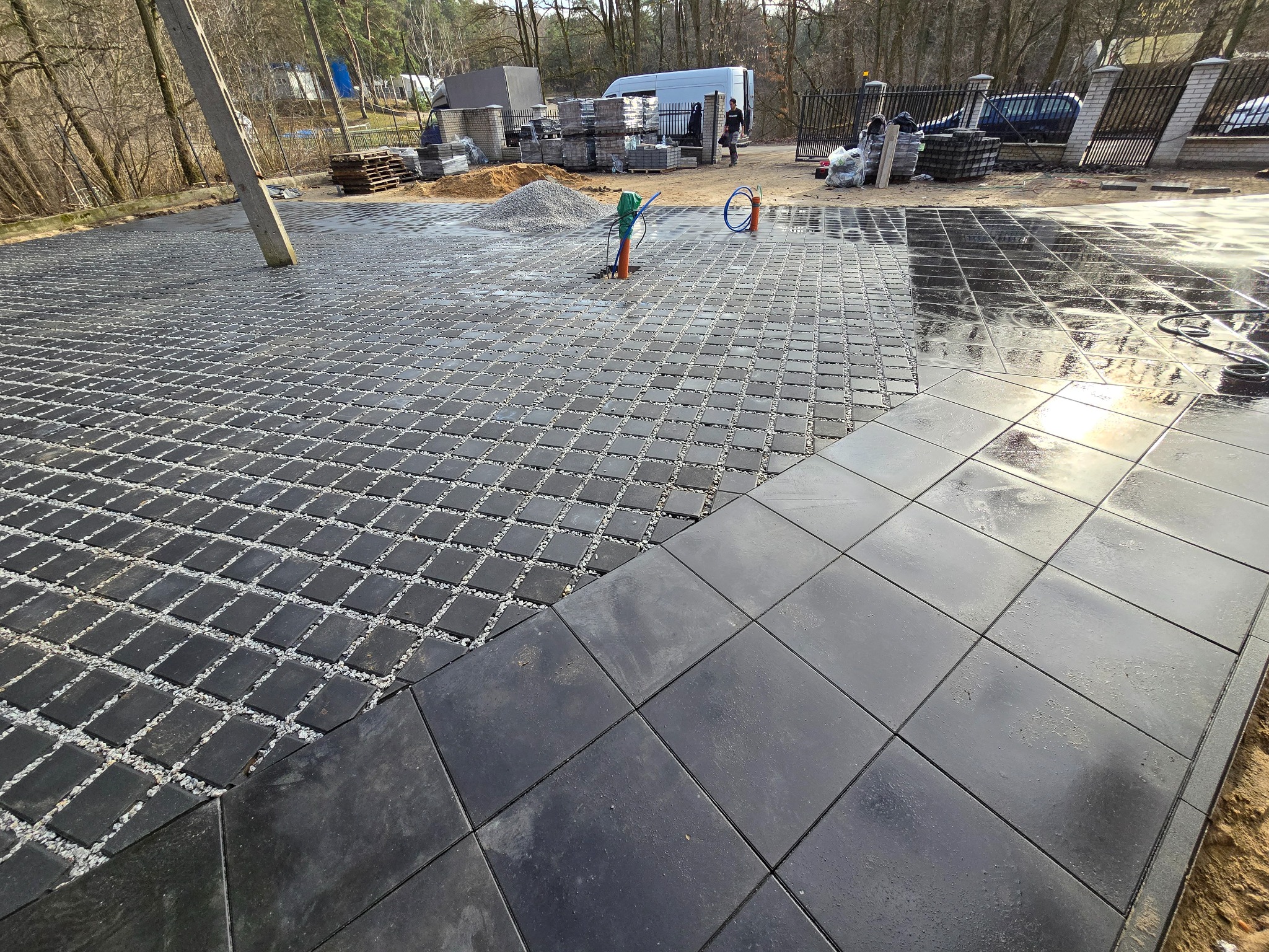 Fragment układanej nawierzchni z dwóch rodzajów kostki: ażurowej wypełnionej grysem i gładkiej, mokrej od wody, z widocznymi elementami instalacji i stosem kostki w tle.