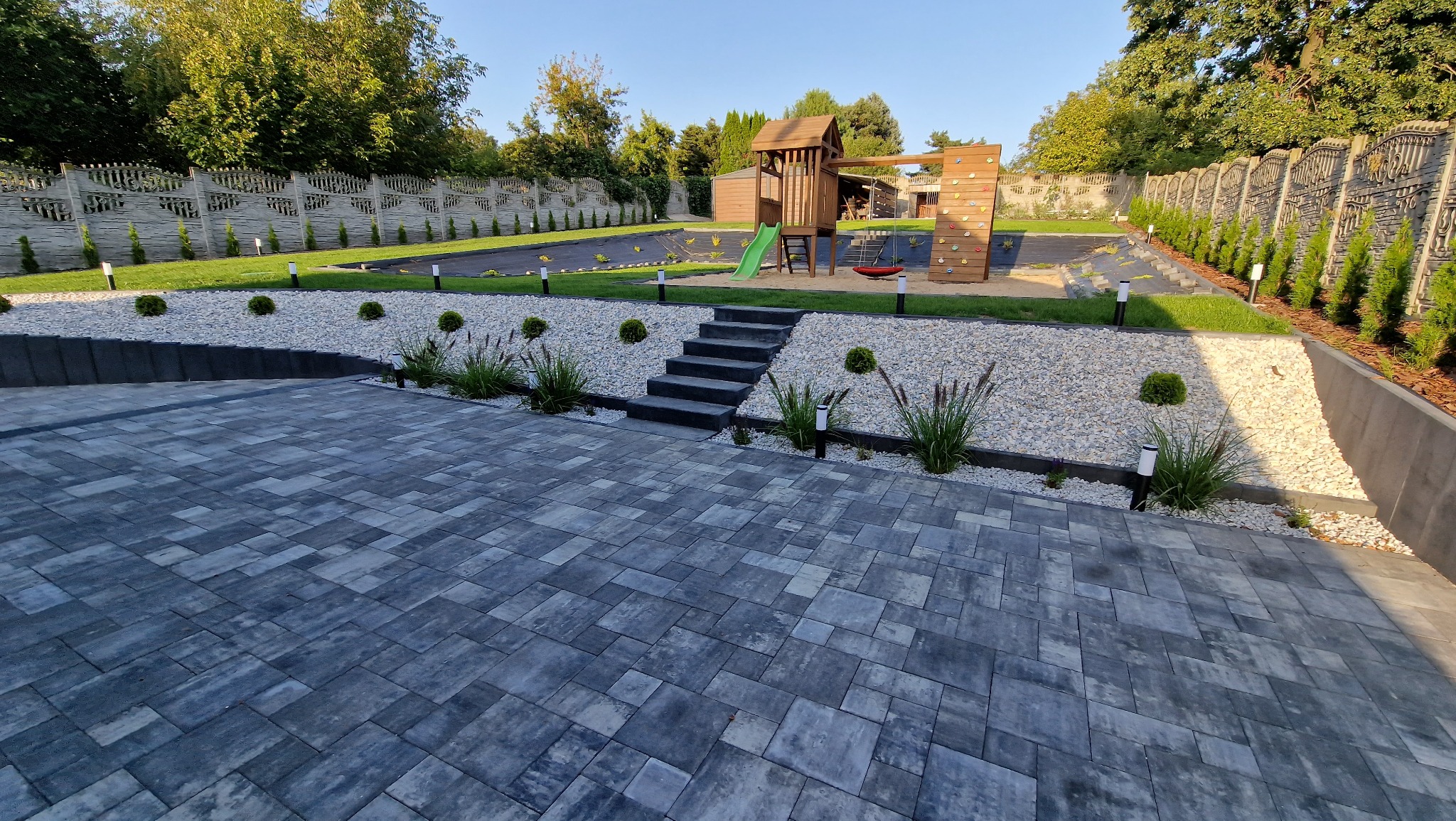 Nowoczesna aranżacja ogrodu z szarej kostki brukowej, schodami prowadzącymi do strefy zabaw dla dzieci z drewnianym placem zabaw, białymi kamieniami i ozdobnymi lampami ogrodowymi.