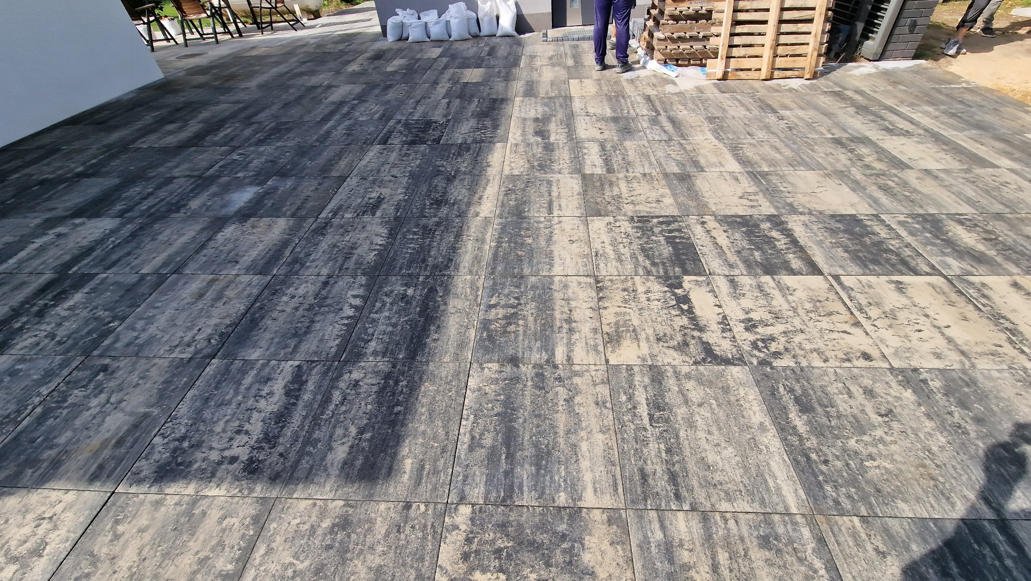 Ułożona nawierzchnia z prostokątnej kostki o nieregularnym wzorze w odcieniach szarości i beżu, z paletą i workami z piaskiem w tle.