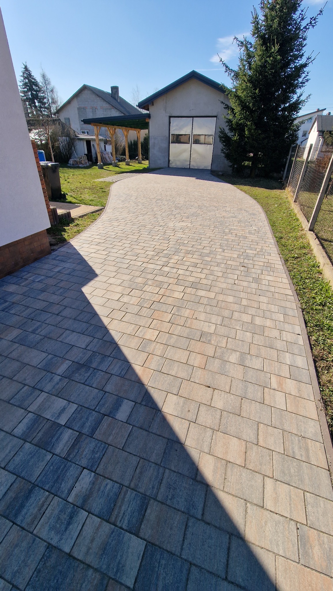 Zakrzywiony podjazd z kostki brukowej w odcieniach szarości i beżu, prowadzący do garażu z metalowymi drzwiami w słoneczny dzień.