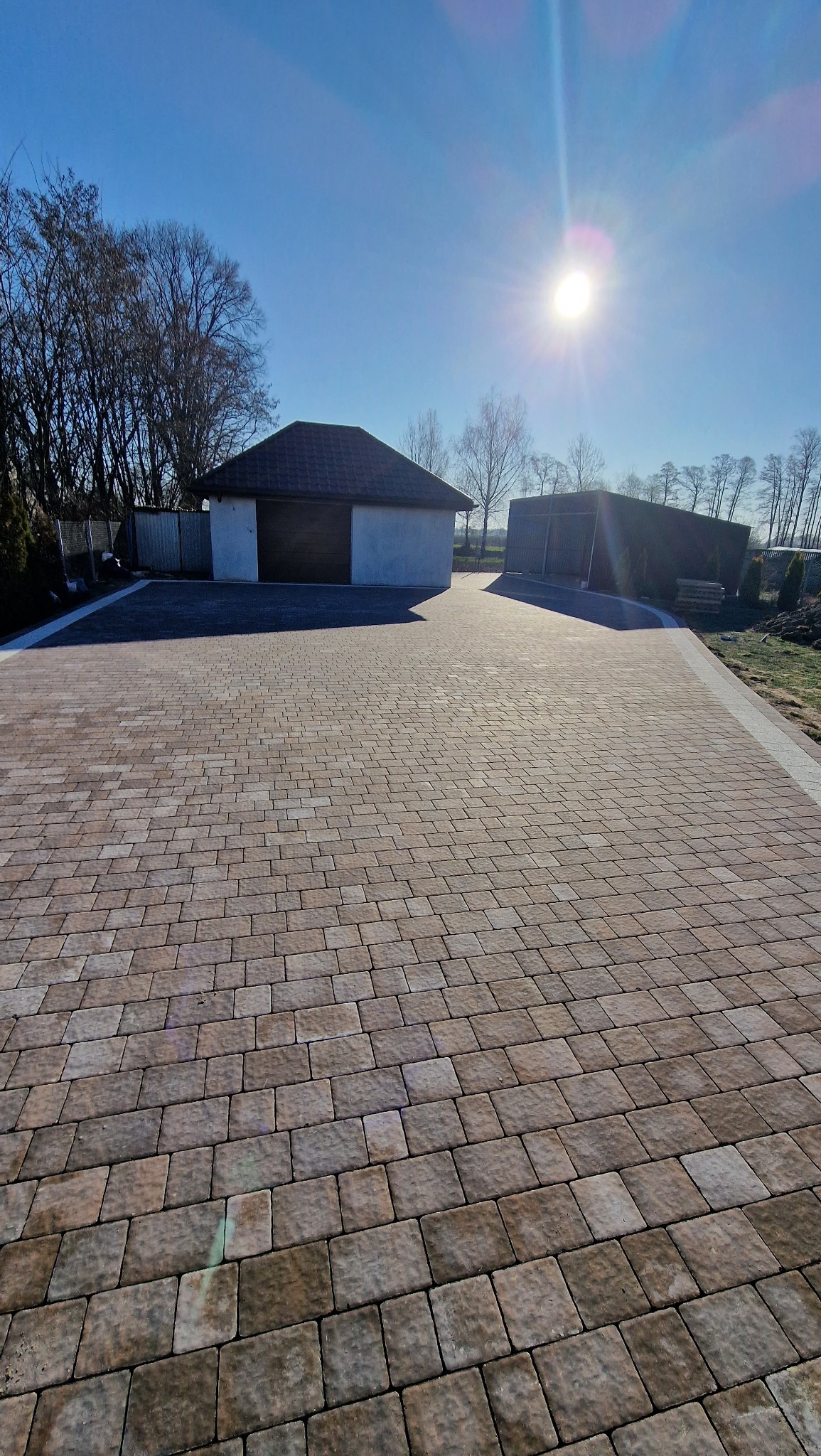 Szeroki podjazd z beżowo-brązowej kostki brukowej prowadzący do garażu z ciemną bramą, w tle drzewa i błękitne niebo z jasnym słońcem.