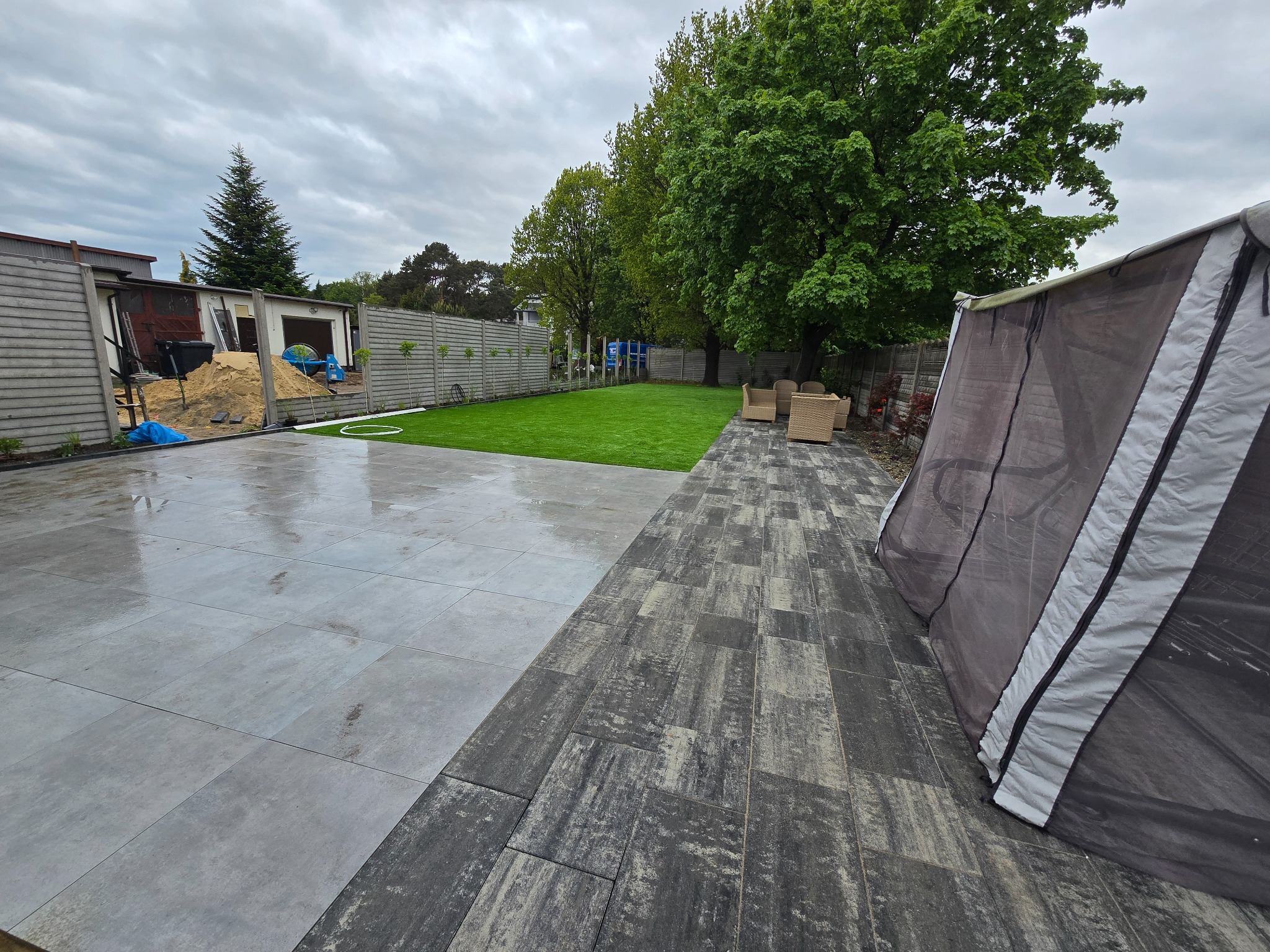 Ułożona, szara kostka brukowa w dwóch wzorach na posesji z trawnikiem i meblami ogrodowymi. Widoczny fragment namiotu i budynek gospodarczy w tle.