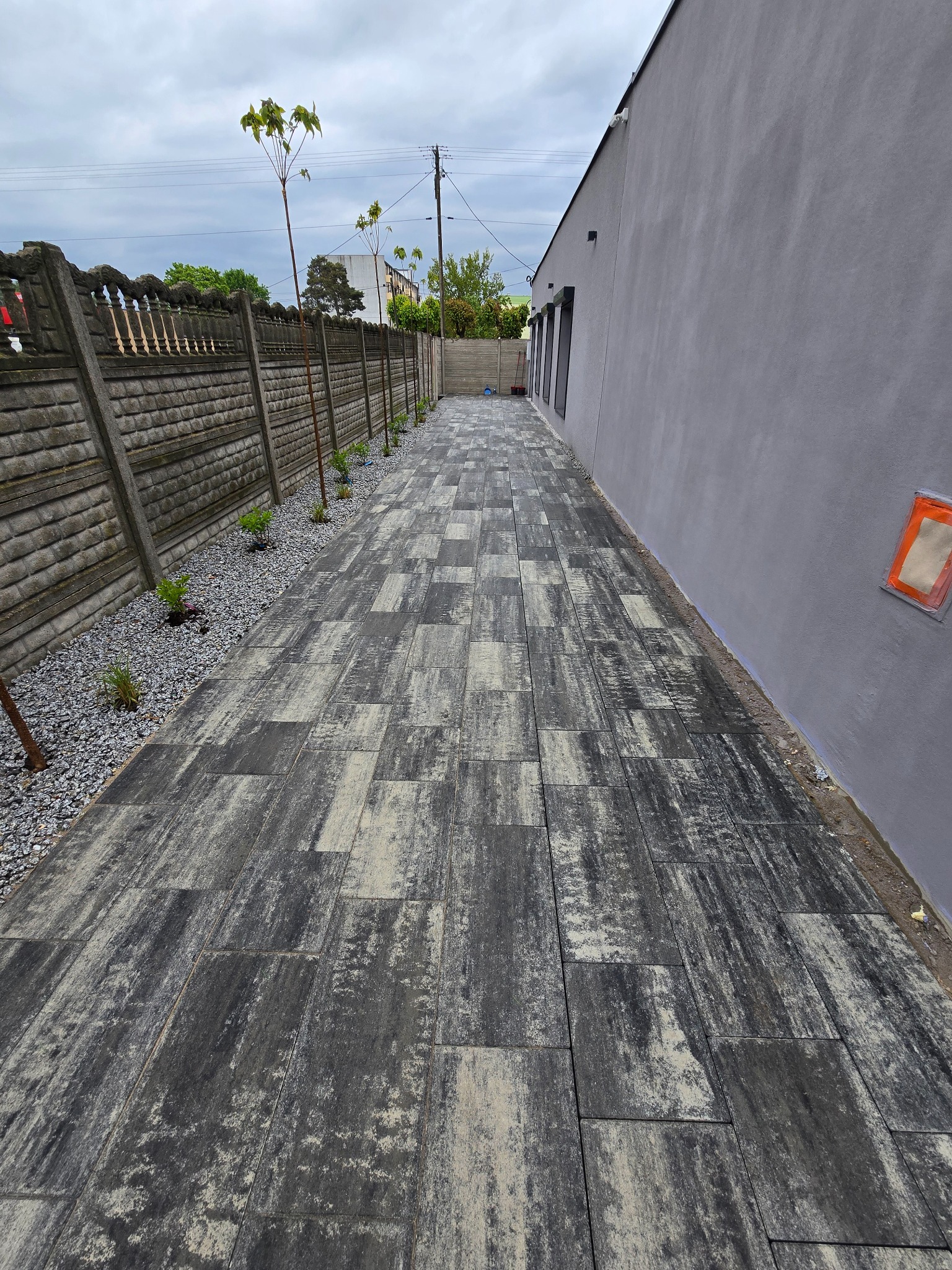 Ułożona, szara kostka brukowa wzdłuż ściany budynku i ogrodzenia. W tle widoczne drzewa i budynek. Ujęcie z perspektywy, podkreślające długość i wzór nawierzchni.