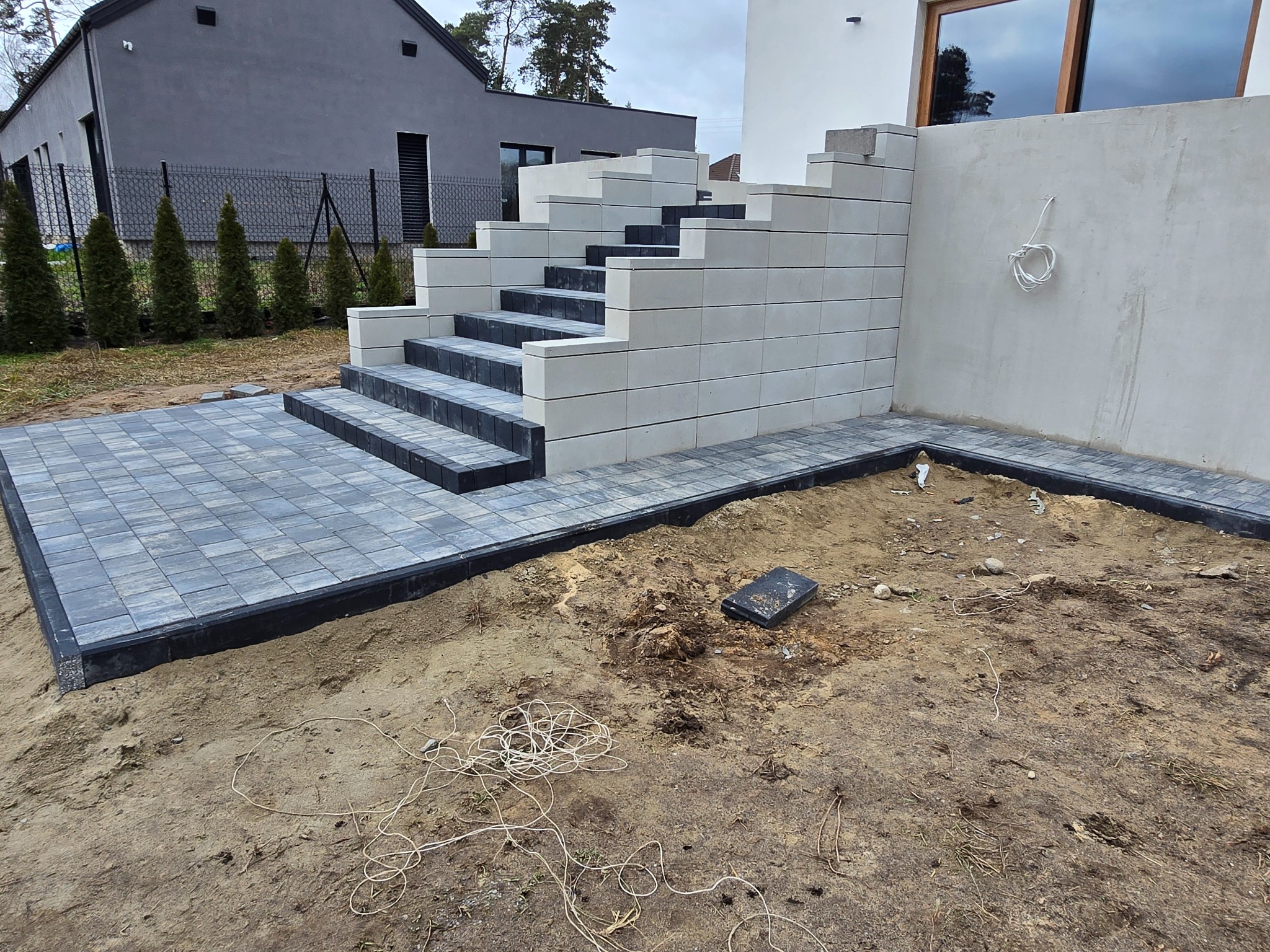 Nowoczesne schody z jasnych, prostokątnych bloków betonowych, prowadzące z szarej kostki brukowej. Widoczne niedokończone prace ziemne i elementy otoczenia.