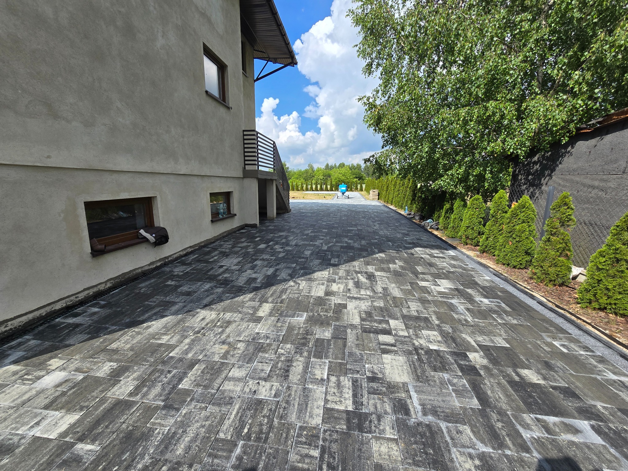 Ułożona, szara nawierzchnia z kostki przy domu z zielonymi tujami wzdłuż ogrodzenia. W tle widoczny błękitny wózek budowlany i drzewa.