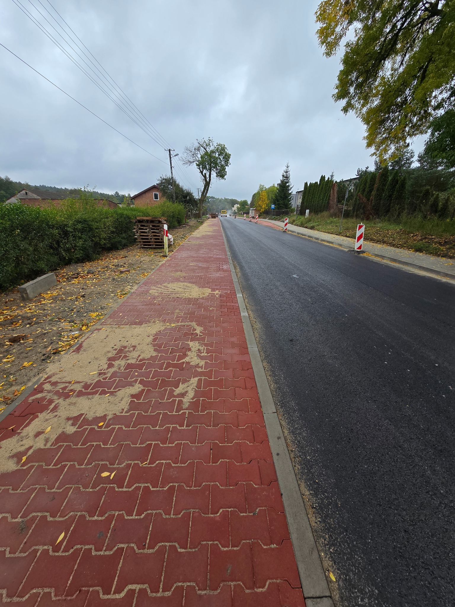 Czerwona kostka brukowa ułożona wzdłuż asfaltowej drogi, widok z perspektywy chodnika. W tle zieleń i budynki, pochmurne niebo. Prace drogowe w toku.