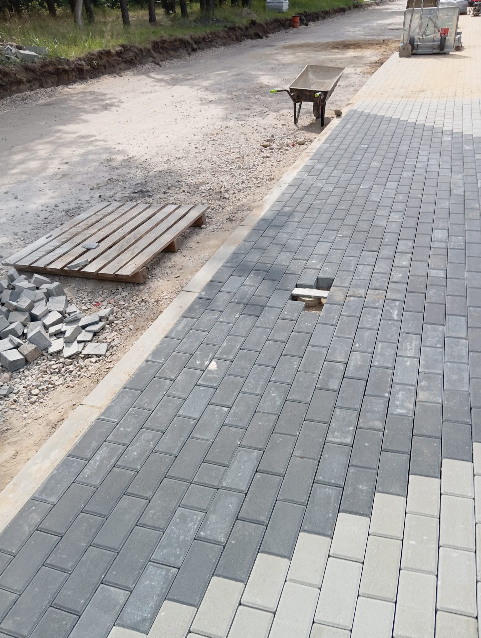 Układanie szarej kostki brukowej w trakcie realizacji. Widoczny fragment z brakiem kostki, paleta, taczka i narzędzia. Częściowo ułożona nawierzchnia.