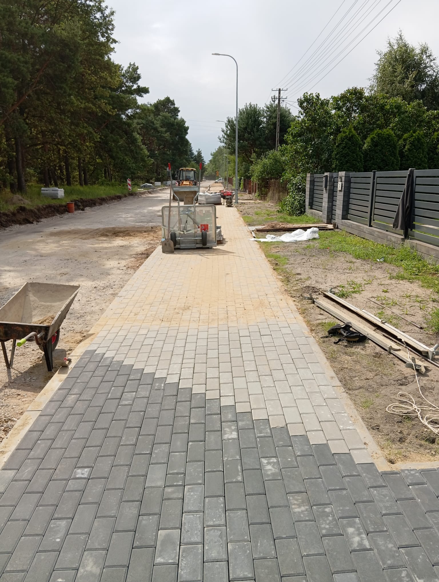 Fragment układanej ścieżki z szarej kostki brukowej, częściowo podsypanej piaskiem. W tle widoczny sprzęt budowlany i teren przygotowany pod dalsze prace.