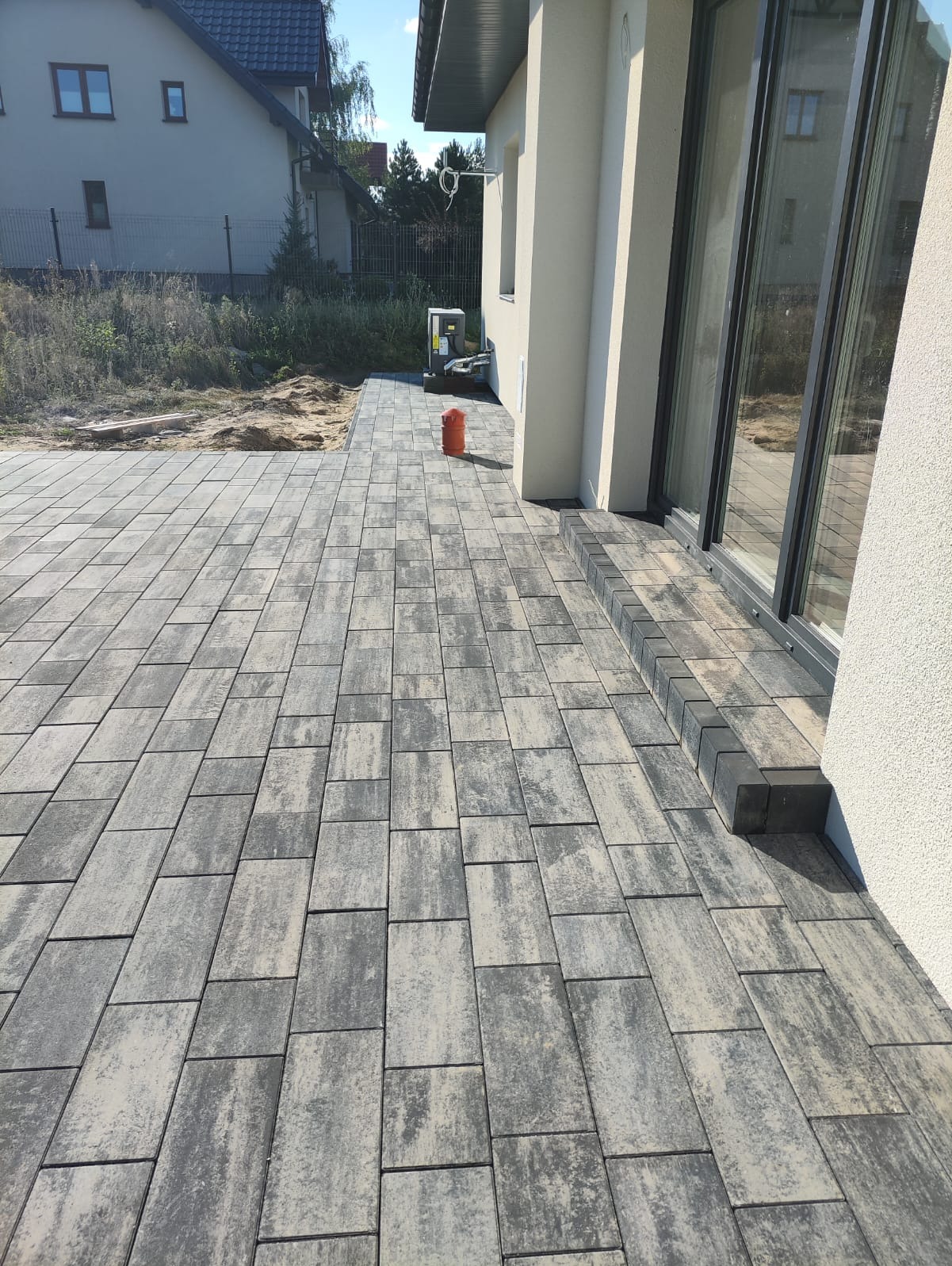 Ułożona szara kostka brukowa na posesji, widok z perspektywy, z elementami wykończeniowymi przy budynku. W tle dom i zieleń. Słoneczny dzień.