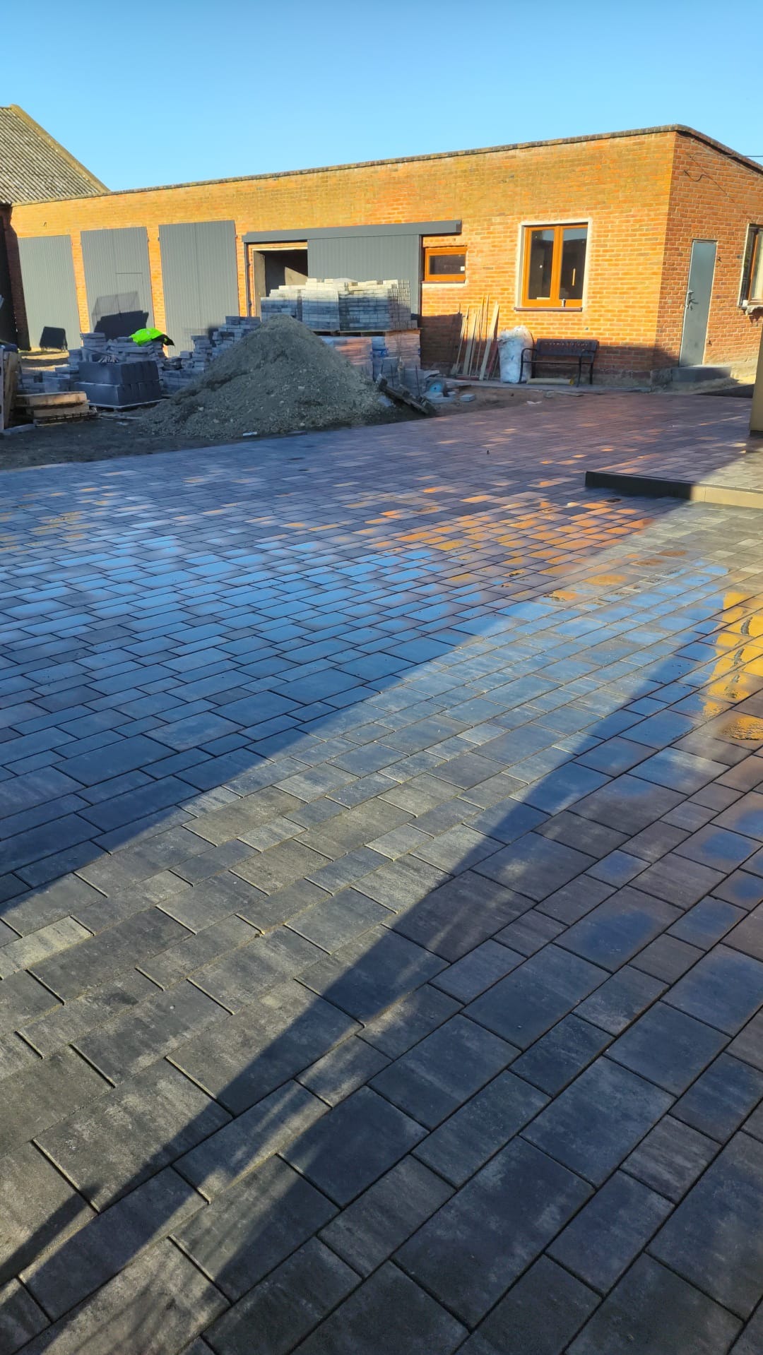 Ułożona, mokra kostka brukowa w odcieniach szarości, z widocznym budynkiem z czerwonej cegły w tle i stertą materiałów budowlanych. Słoneczny dzień.