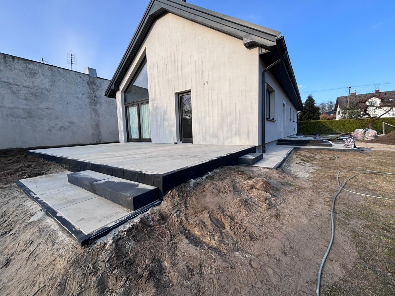 Nowoczesny taras z jasnych płyt, obrzeża z ciemnej kostki, schody. Dom z jasną elewacją w tle, świeżo ułożona ziemia wokół. Minimalistyczny design.