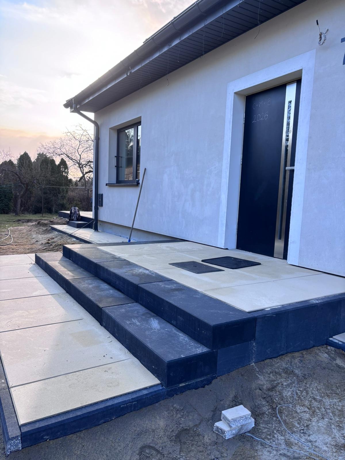 Wykończone schody wejściowe z kostki betonowej w odcieniach szarości i beżu, prowadzące do domu z czarnymi drzwiami. Widoczne detale wykończenia i otoczenie domu.