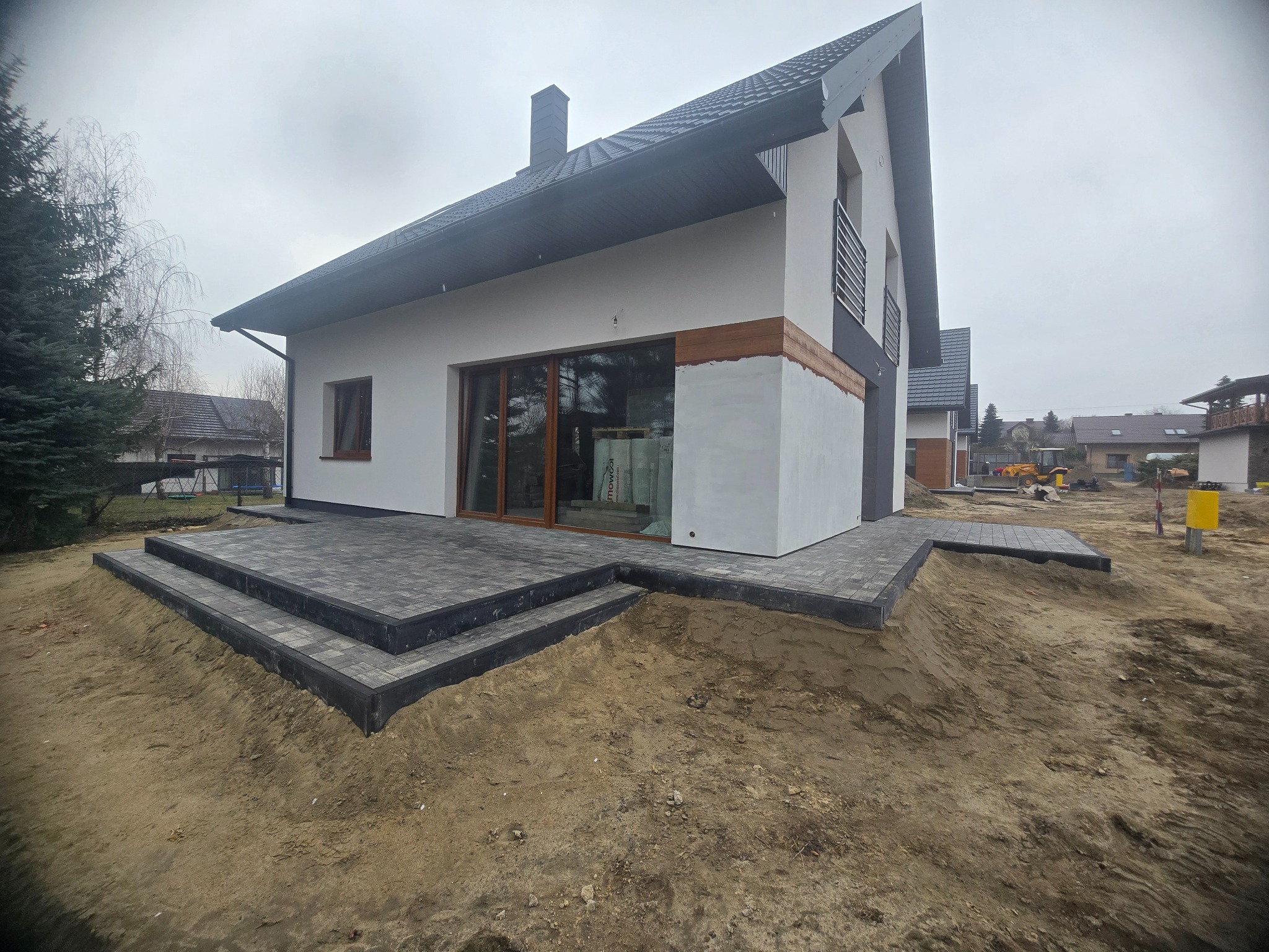 Nowoczesny dom z szarą kostką brukową na tarasie i schodach wejściowych. Kostka ułożona na ziemi, widok na elewację budynku i fragment ogrodu. Pochmurny dzień.