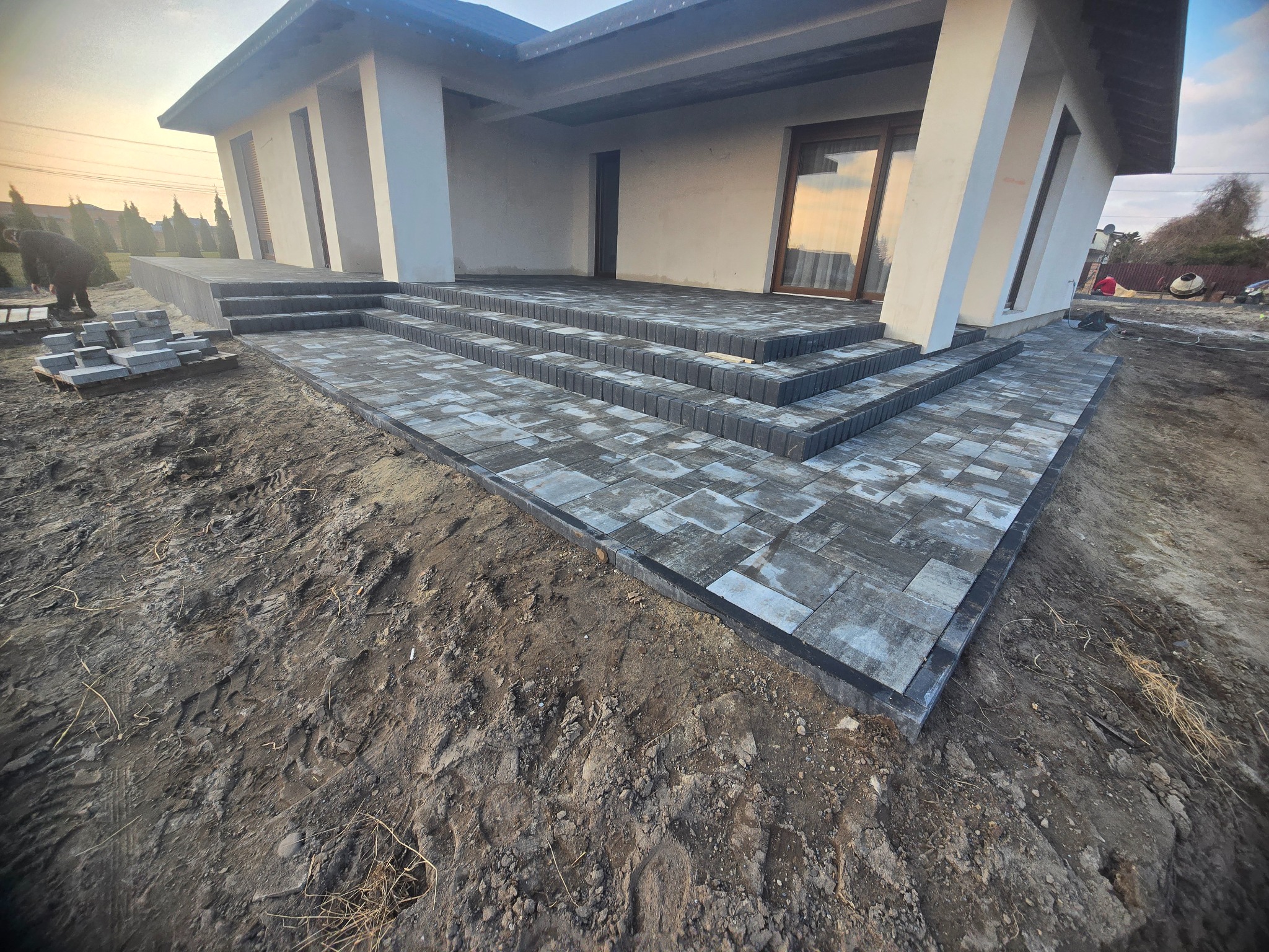 Nowoczesny dom z tarasem i schodami wyłożonymi szarą kostką. Częściowo ułożona nawierzchnia, paleta z kostką w tle. Widok z dołu, podkreślający geometrię.