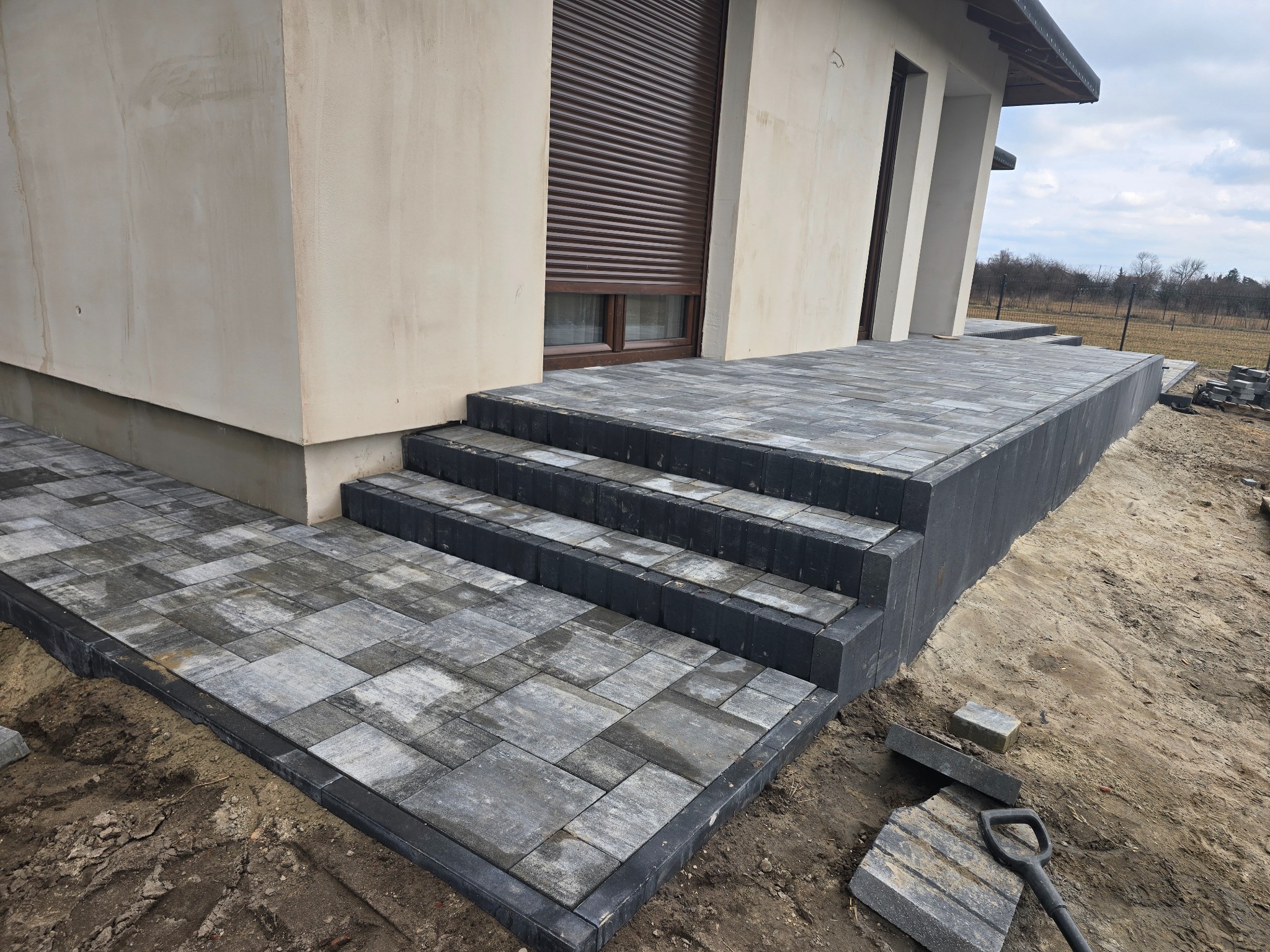 Schody i chodnik z szarej kostki brukowej prowadzące do domu z brązowymi roletami, widoczne narzędzia i niedokończone prace ziemne.