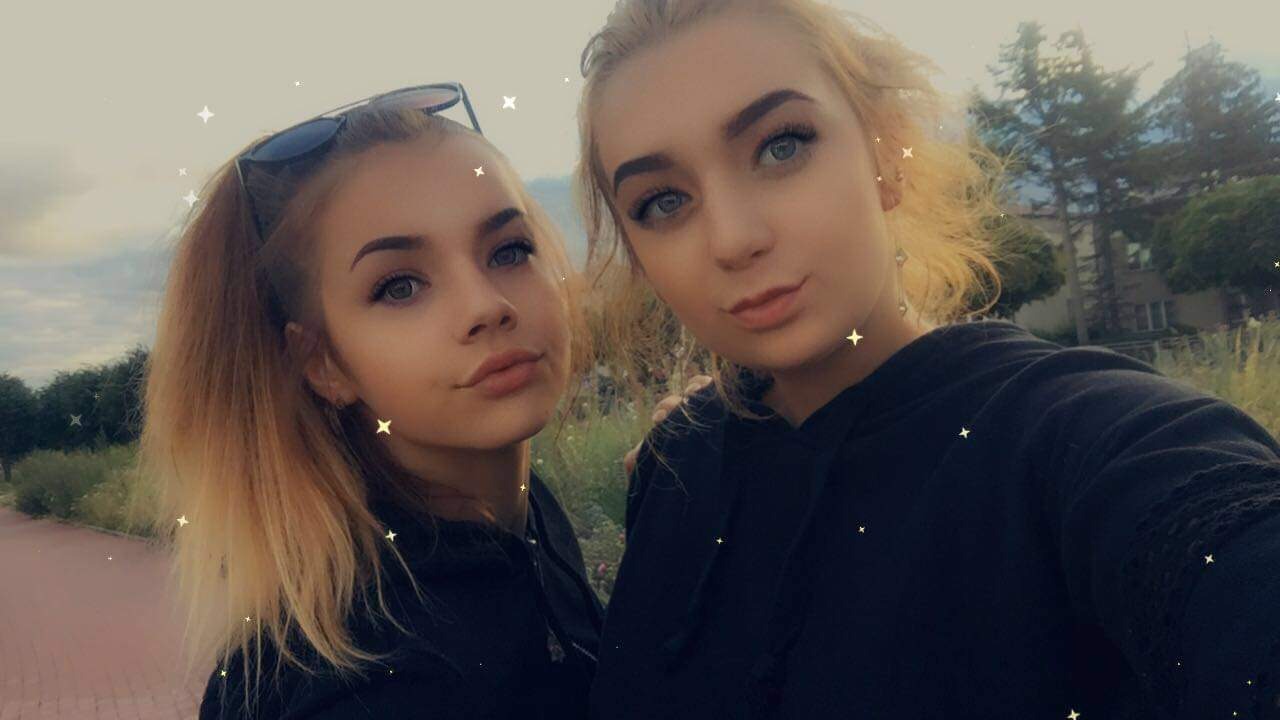 Dwie młode kobiety w Mławie, uśmiechnięte, pozują na tle zieleni i chodnika, z efektami specjalnymi w postaci błyszczących gwiazdek.