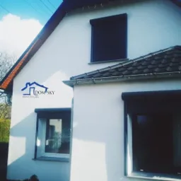 Biały dom jednorodzinny z trzema oknami z roletami zewnętrznymi, widoczne logo firmy na ścianie, błękitne niebo w tle.