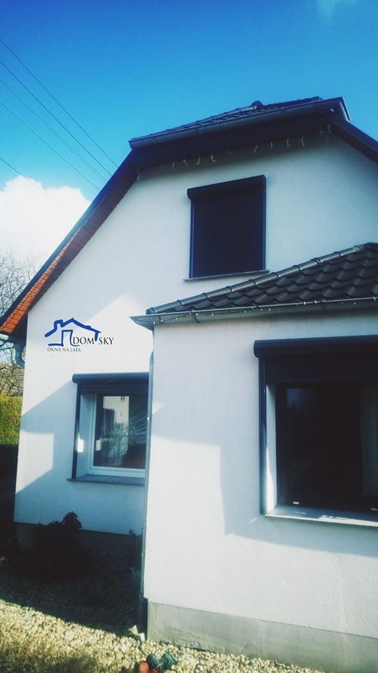 Biały dom jednorodzinny z trzema oknami z roletami zewnętrznymi, widoczne logo firmy na ścianie, błękitne niebo w tle.