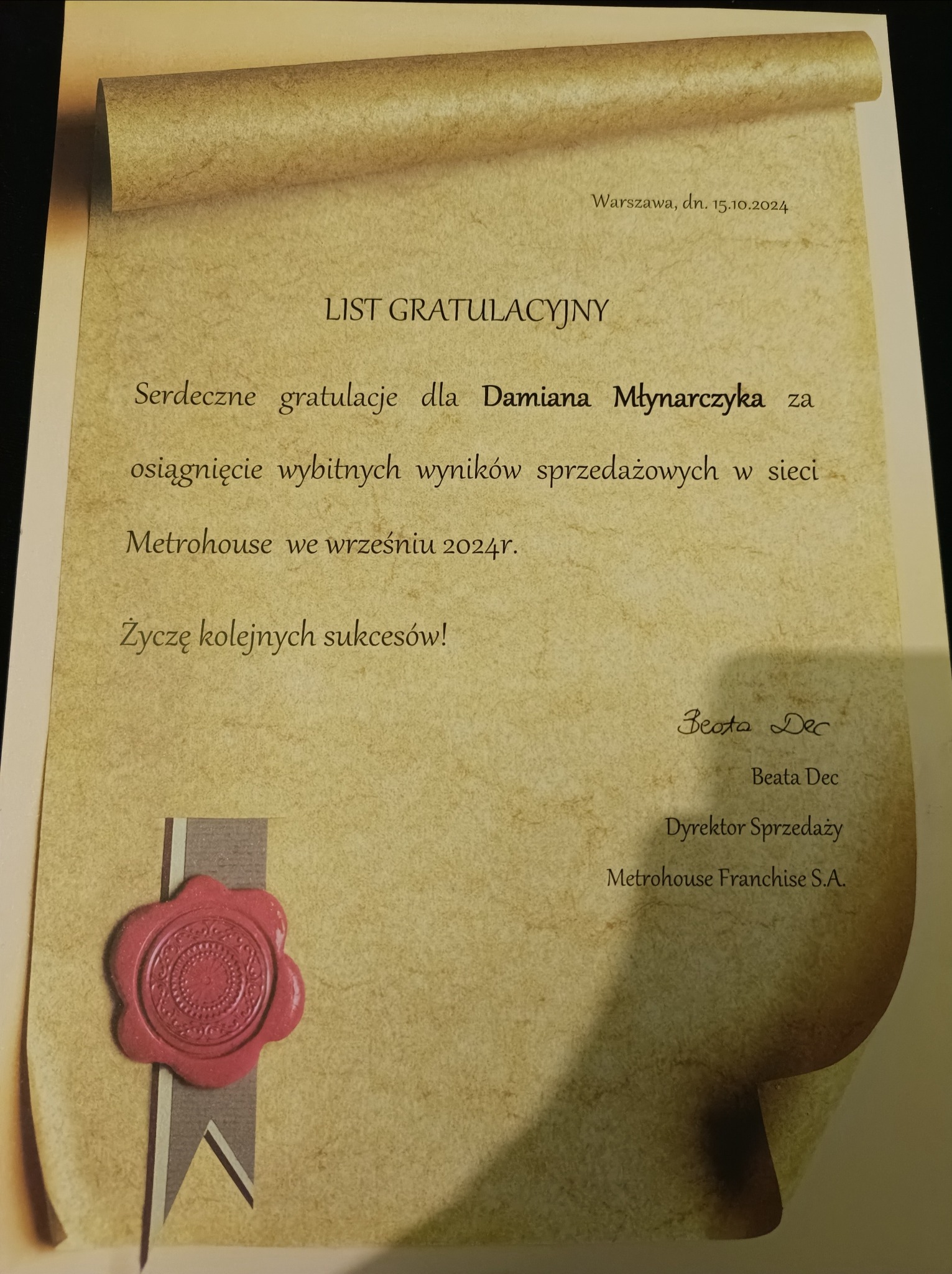 Uroczysty list gratulacyjny dla Damiana Młynarczyka za wybitne wyniki sprzedażowe w sieci Metrohouse, opatrzony czerwoną lakową pieczęcią i podpisem Dyrektora Sprzedaży Beaty Dec.