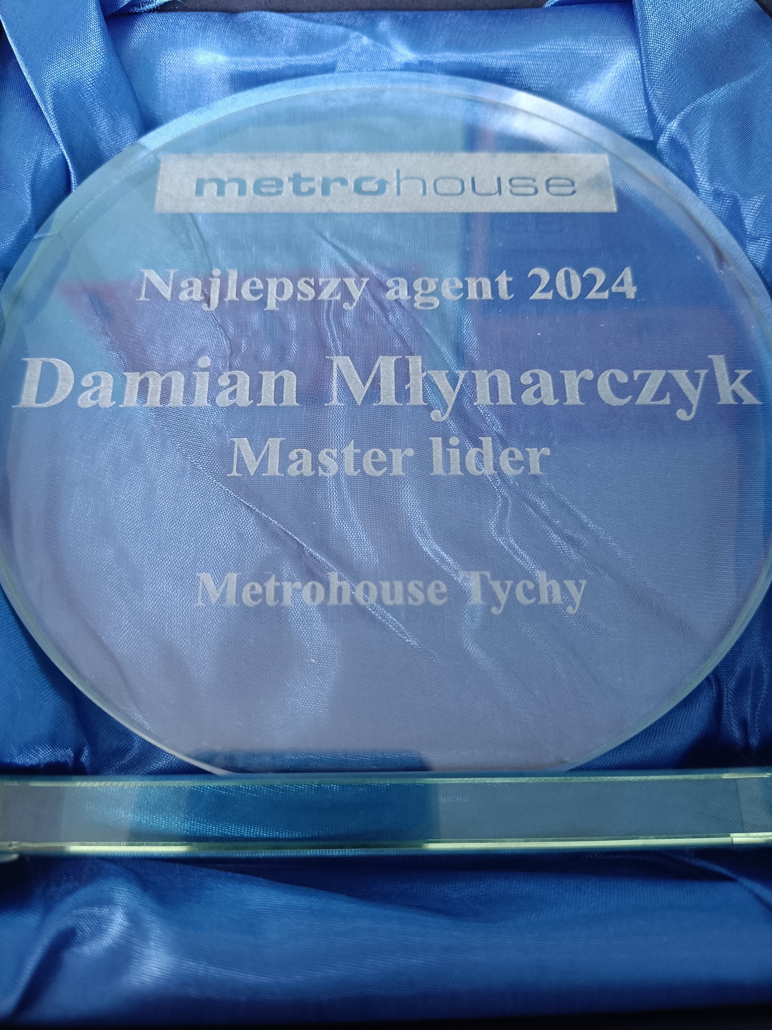 Szklana statuetka z grawerem 'Najlepszy agent 2024 Damian Młynarczyk Master lider Metrohouse Tychy' na niebieskim, satynowym tle.