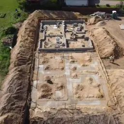 Widok z drona na plac budowy z wykopami pod fundamenty i częściowo wylane fundamenty budynku, widoczne szalunki i pracownicy budowlani.