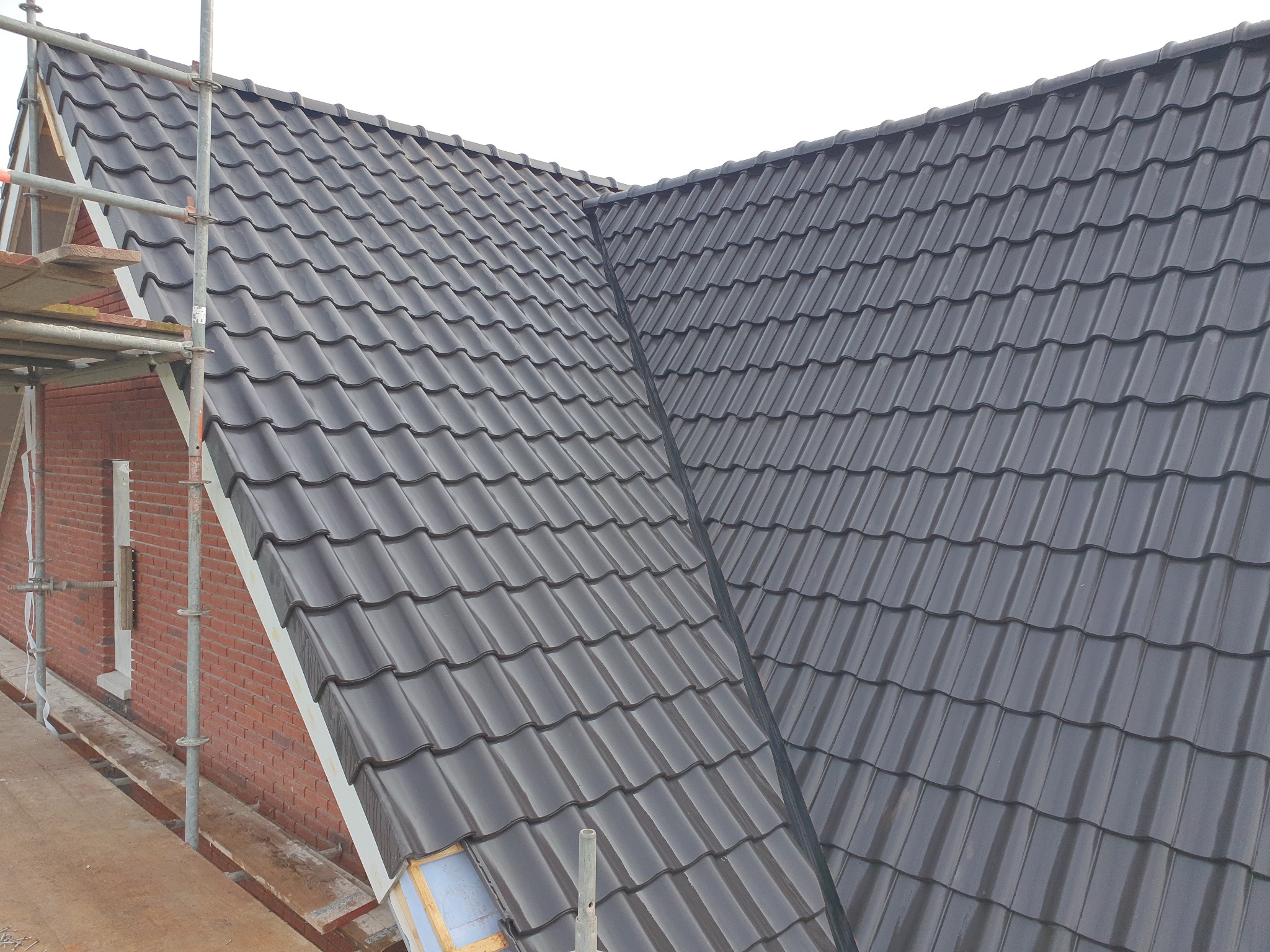 Nowy, błyszczący dach z ciemnoszarej dachówki ceramicznej, widok z rusztowania, fragment muru z czerwonej cegły w tle.