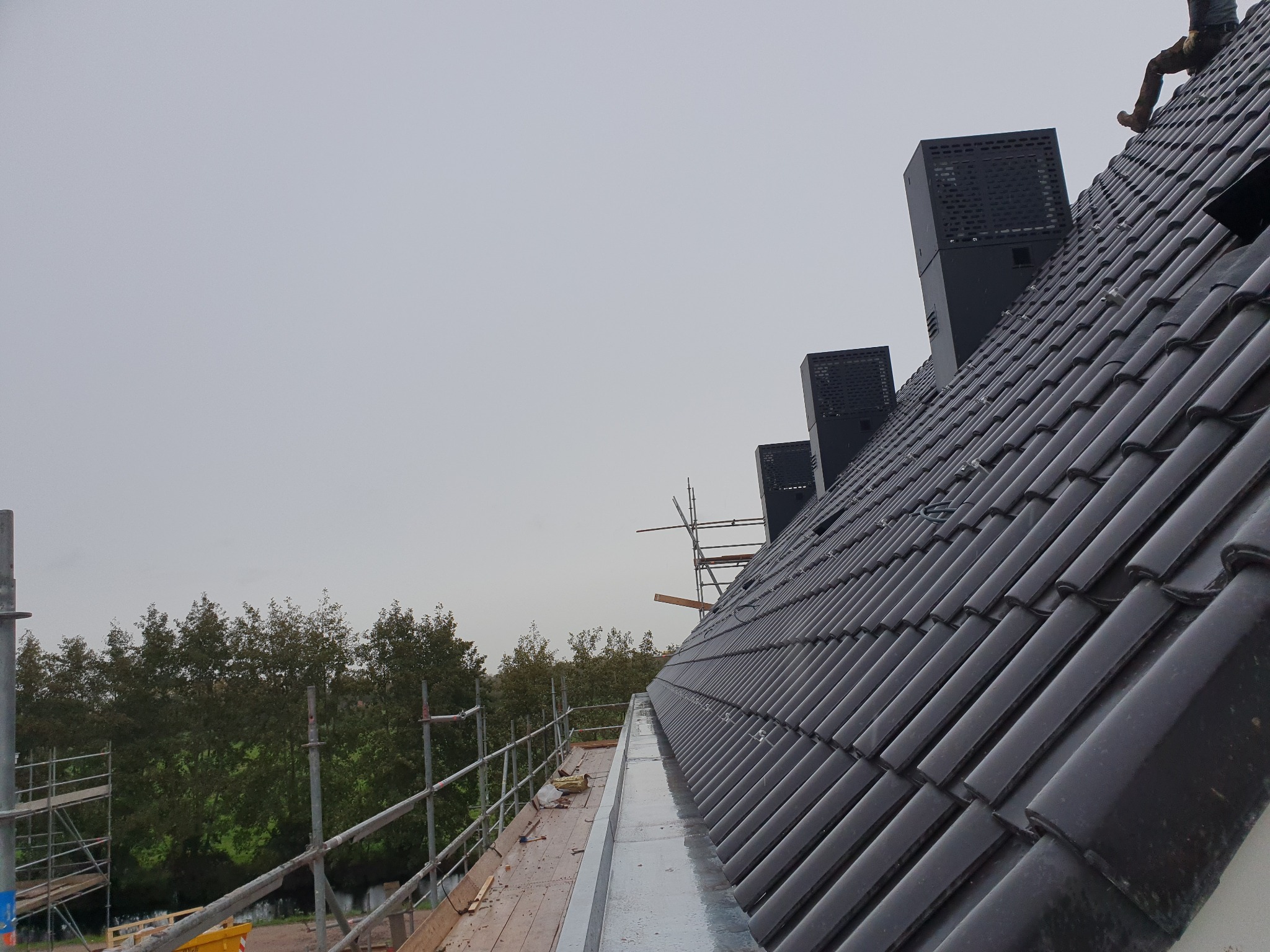 Nowy, czarny dach z dachówki ceramicznej, z widocznymi kominami wentylacyjnymi, zamontowany na tle szarego nieba i zielonych drzew, częściowo widoczne rusztowanie.