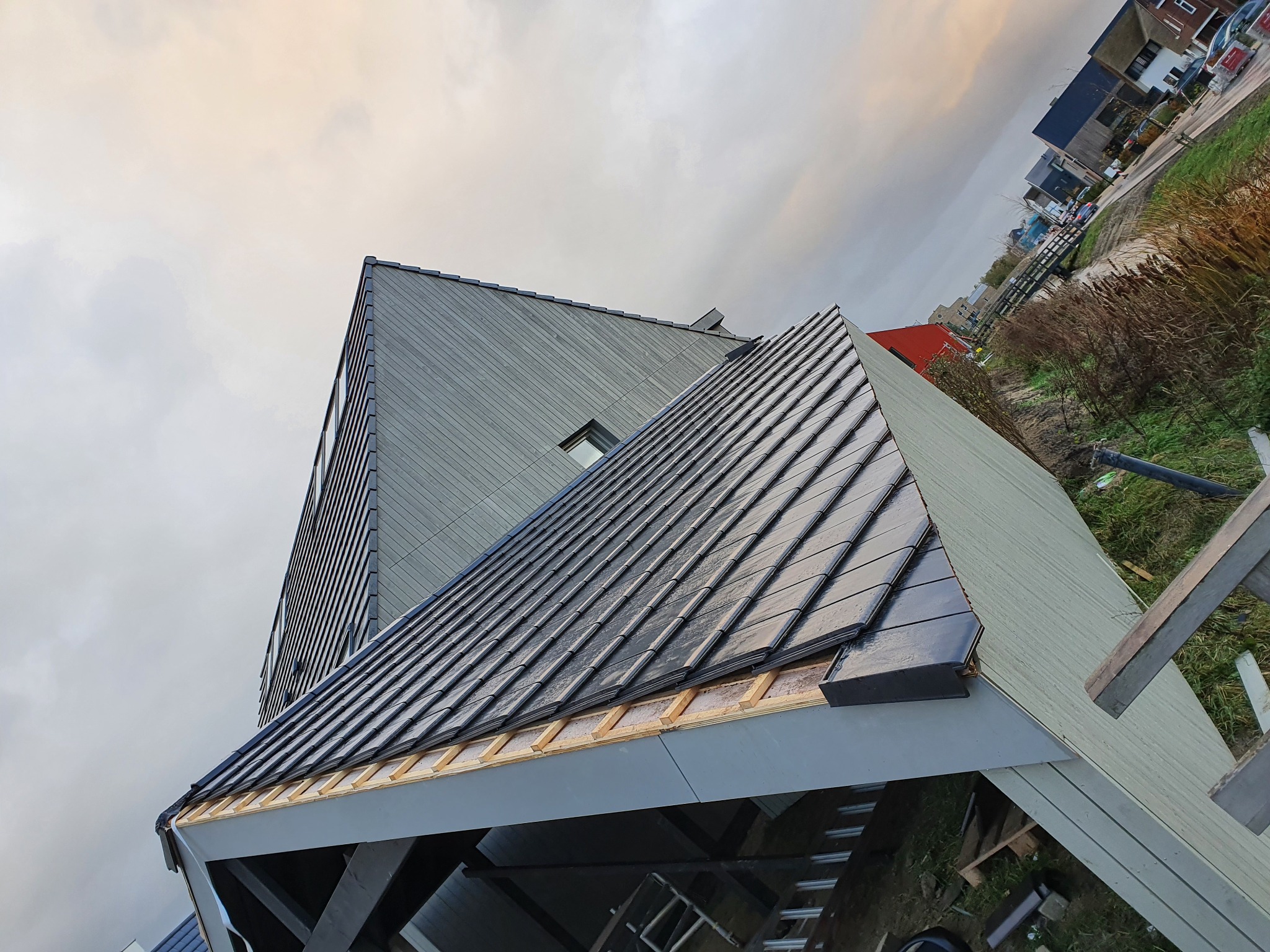 Nowy dach z ciemnej dachówki ceramicznej na tle nowoczesnego domu z szarą elewacją, widoczne drewniane elementy konstrukcji dachu i fragment drabiny.