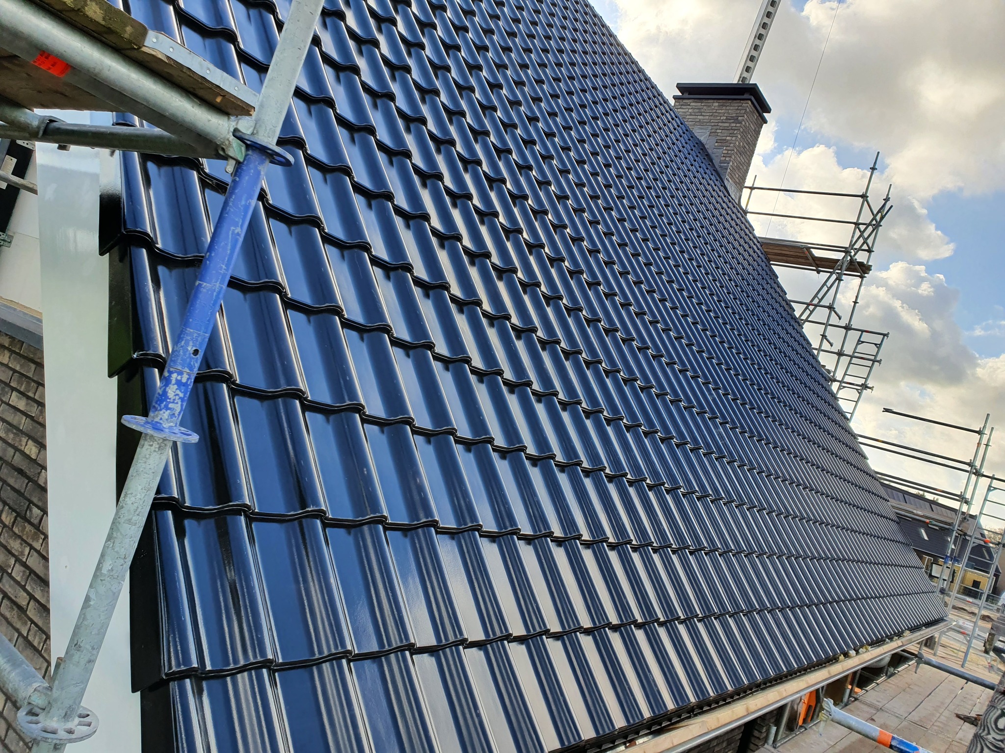 Błyszczący, czarny dach z dachówki ceramicznej widziany z rusztowania, komin w tle, częściowo zachmurzone niebo.