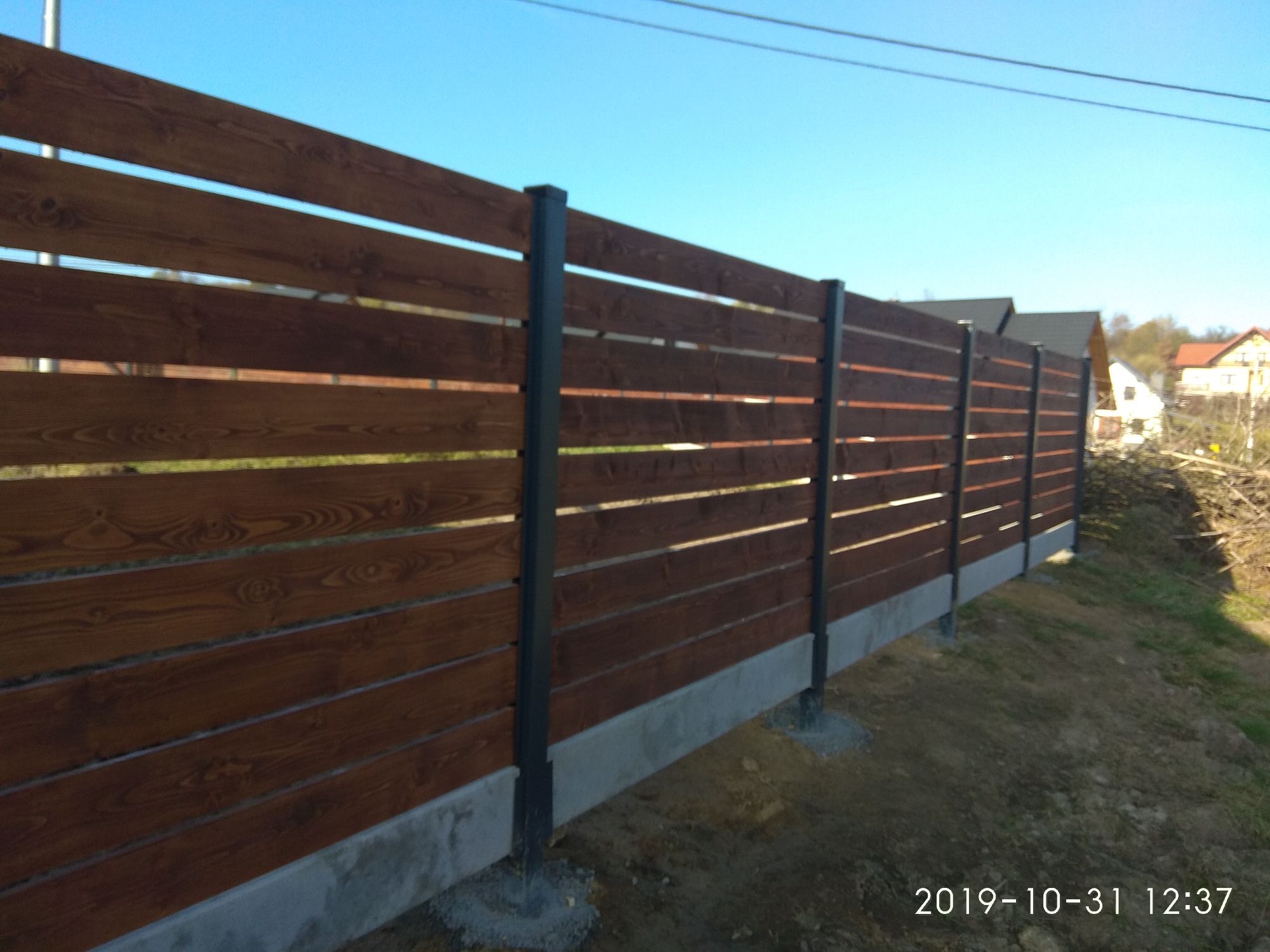 Poziome ogrodzenie z desek w kolorze ciemnego drewna, wsparte na grafitowych słupkach, osadzone na betonowej podmurówce, widoczne z boku na tle błękitnego nieba i zabudowań.