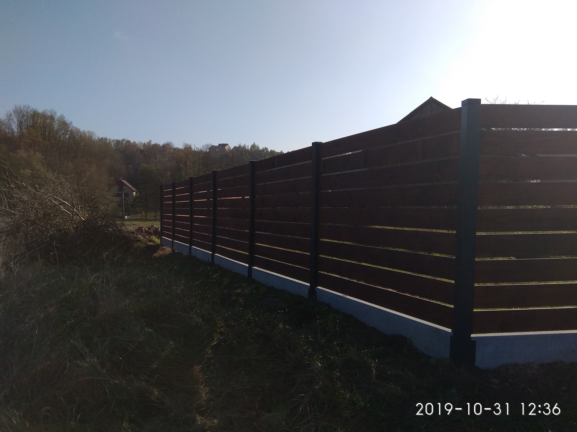 Długie, poziome ogrodzenie drewniane w kolorze ciemnego brązu, z szarymi słupkami i podmurówką, na tle zielonego trawnika i drzew w oddali.