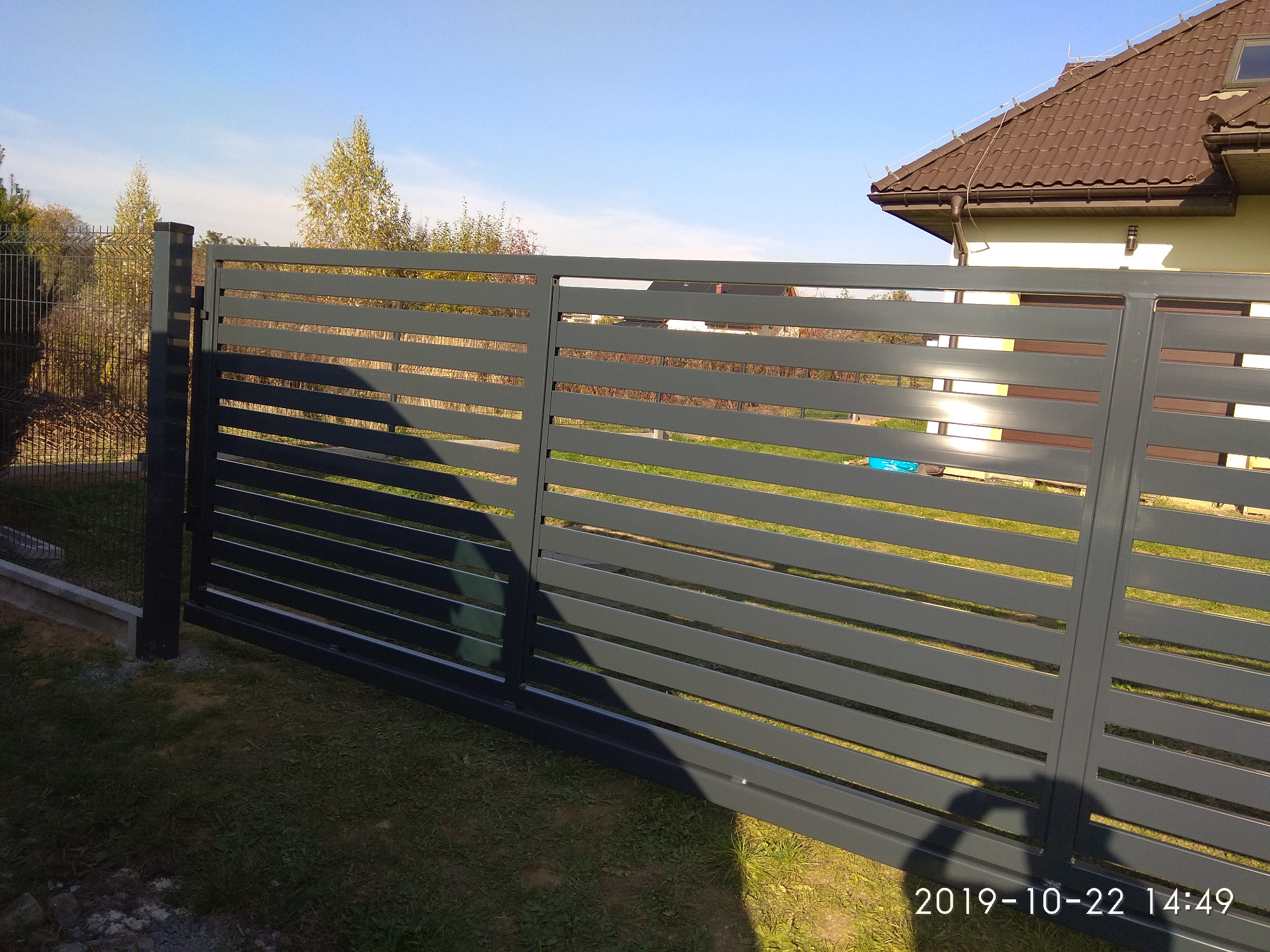 Nowoczesna, przesuwna brama wjazdowa z poziomych, szarych profili, widoczna na tle domu i siatki ogrodzeniowej, z datą wykonania zdjęcia w prawym dolnym rogu.