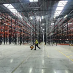 Wnętrze hali magazynowej z wysokimi, metalowymi regałami w kolorze czarnym i pomarańczowym; pracownik w odblaskowej kamizelce ciągnie wózek z nawiniętą taśmą.